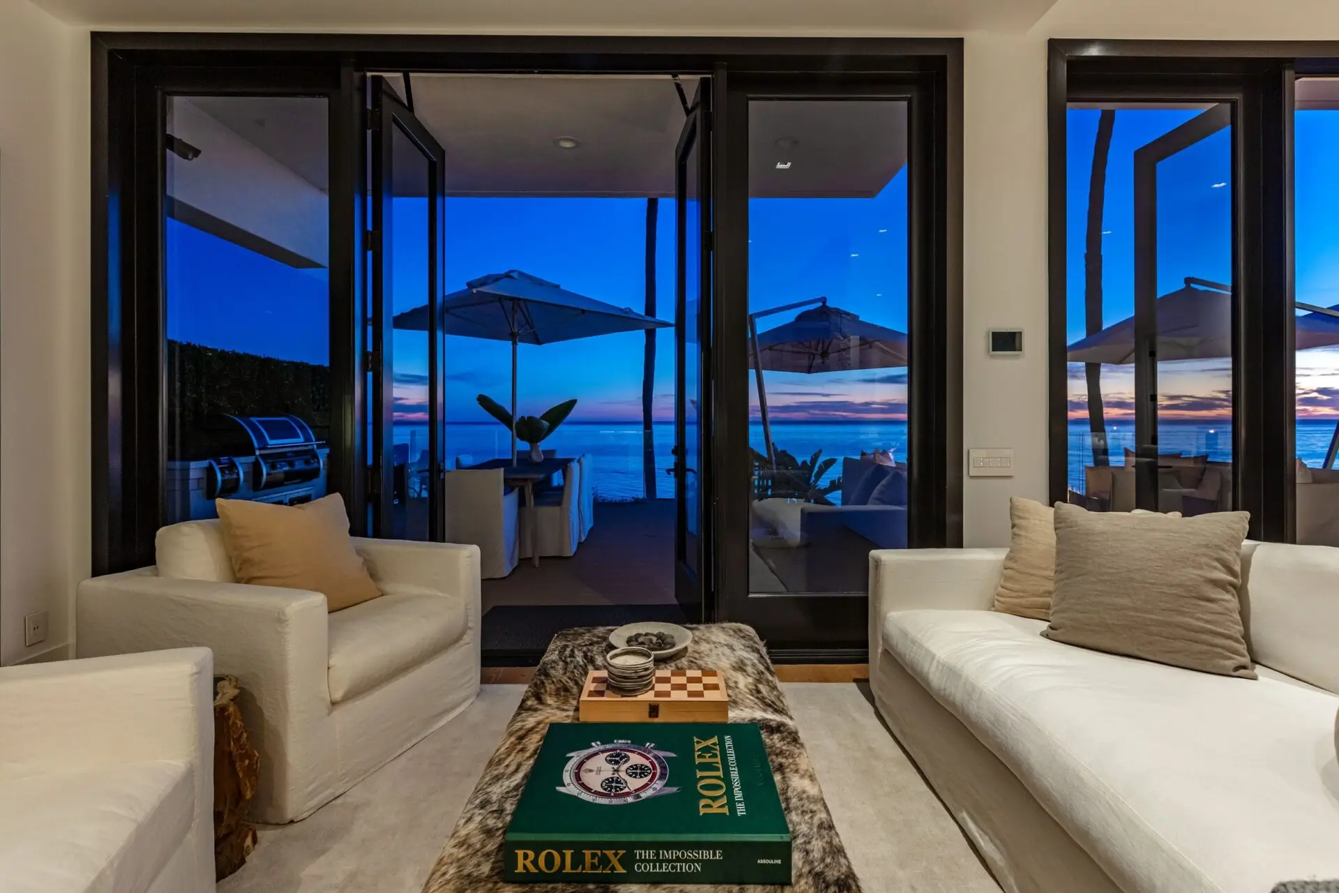 A sitting area at Carrara's Malibu house at dusk, two white armchairs, a chess set, and deck umbrellas beyond