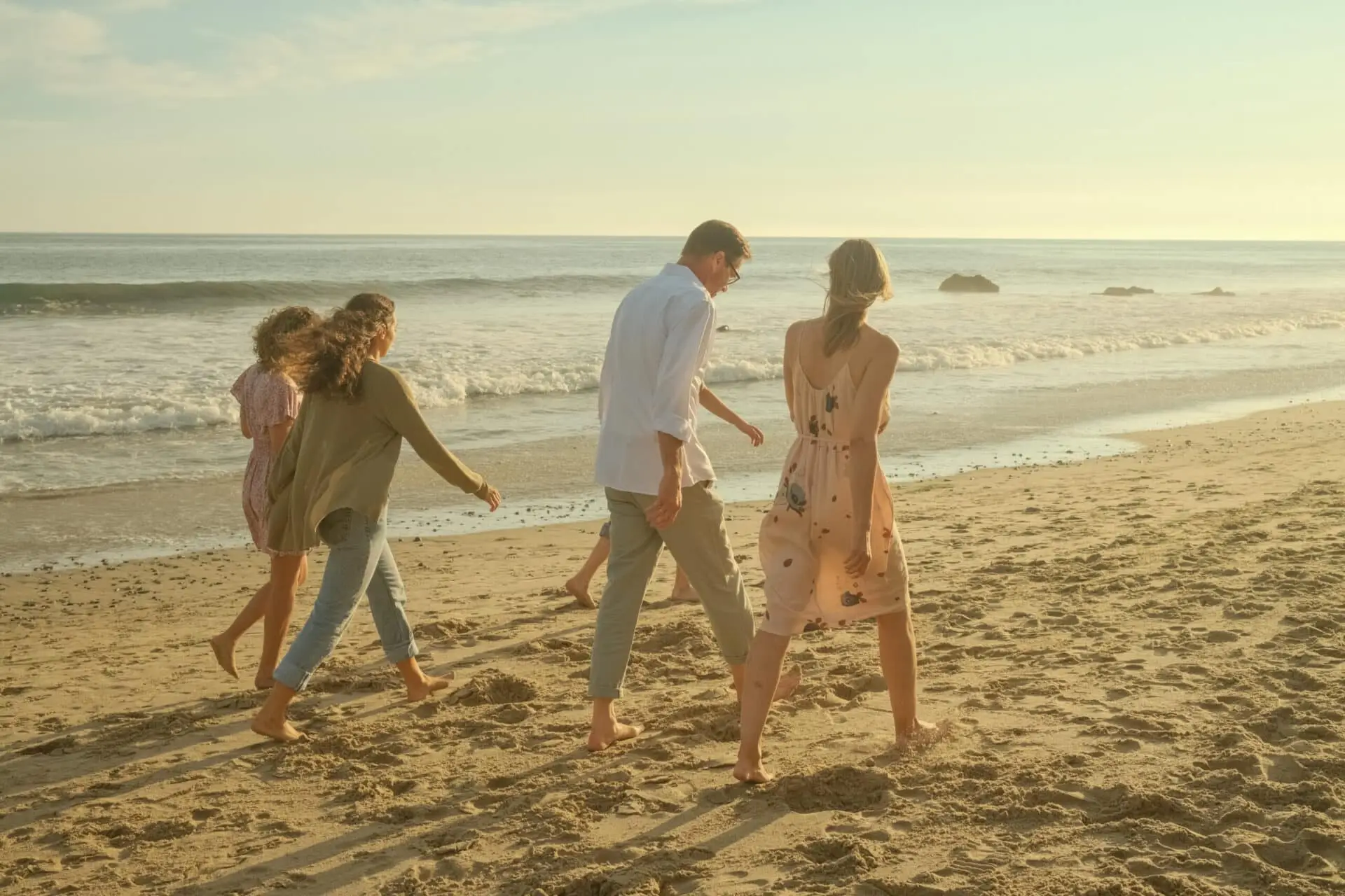 A family of five walking barefoot along the surf line at Malibu in low golden light, faces turned away