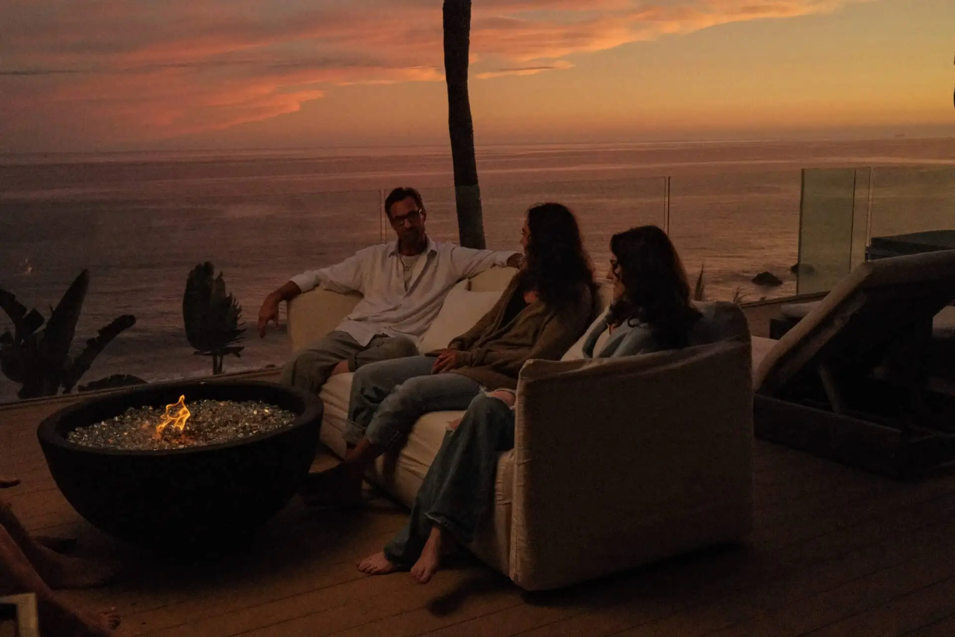 Three people talking around a fire bowl on Carrara's Malibu deck at sunset, the ocean behind the glass rail