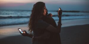 Couple embracing on beach at twilight, each using their own smartphone against ocean backdrop.