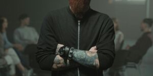 Man with beard and tattoos wearing black jacket, arms crossed, showing wristwatch. People visible in background.