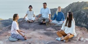 Group meditating on rocky cliff overlooking ocean, diverse people in casual clothes practicing mindfulness outdoors.