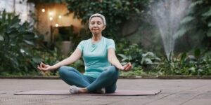 Mature woman with short gray hair meditating outdoors on yoga mat, surrounded by plants and string lights.