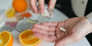 Hands dispensing medication pill from bottle near orange slices and blister packs.