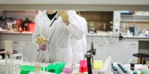 Scientist in lab coat conducting experiment with purple liquid in flask, surrounded by lab equipment.
