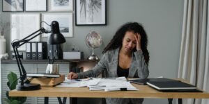 Woman looking stressed while working at desk with papers, laptop, and office decor around her.