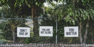 Three white signs on a chain-link fence with encouraging messages: "DON'T GIVE UP", "YOU ARE NOT ALONE", and "YOU MATTER".