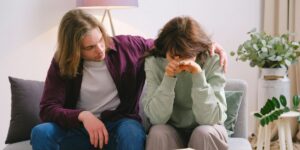 Two people on couch; one comforting the other who appears distressed, covering face.