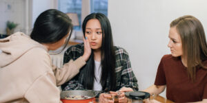 Alt Text: Three young women at table during intervention, one comforting distressed friend as another looks on.