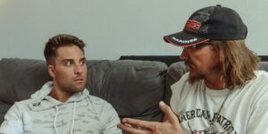 Two men on couch, one looking surprised as the other gestures while speaking, wearing Marines cap.