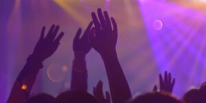 Silhouettes of raised hands at a concert or music festival. The background is lit with vibrant purple and orange lights, creating a dreamy, atmospheric effect. Lens flares and light orbs add to the energetic ambiance.