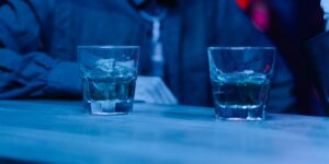 Two glasses of clear liquor with ice on a table, bathed in blue nightclub lighting.