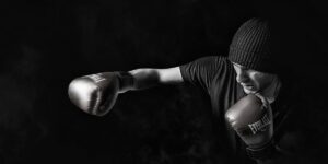 This image shows a boxer in a dark, dramatic lighting. The boxer is wearing boxing gloves and a beanie, with their fist raised, suggesting a powerful, intense, and focused moment in a training or competition setting.