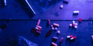 Assorted pills and capsules scattered on a dark wooden surface under blue lighting.