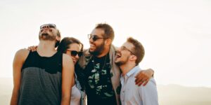 Group of four young adults laughing together outdoors, wearing sunglasses, with sunlit mountains in background.