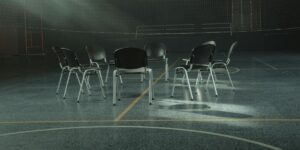 Circle of empty chairs on a dimly lit indoor sports court, suggesting a group therapy or support meeting setting.