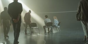 People gathering around chairs in a dimly lit room with dramatic light beams, suggesting a support group or therapy session setting