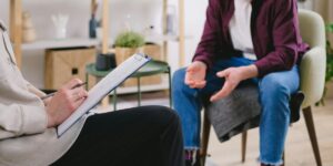 Therapist taking notes while patient in purple sweater gestures during counseling session in modern office