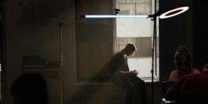 This image shows a person sitting alone in a dimly lit room, focused on their smartphone. The lighting is dramatic, with a backlit window and a bright, halo-like studio light overhead, creating a pensive, contemplative atmosphere.