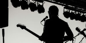 Silhouette of a musician holding a guitar and standing at a microphone on stage, with stage lights visible above