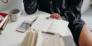 Person in black shirt writing in notebook at white desk, with smartphone and coffee cup nearby.