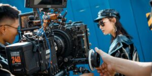 The image shows a movie production scene, with a camera crew setting up and preparing a shot. It features a complex professional camera rig with various equipment, and a crew member, likely the director or cinematographer, adjusting and setting up the camera.