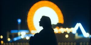 Silhouette of a person against a bright orange Ferris wheel at night, with colorful carnival lights in the background.