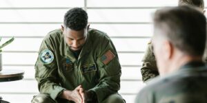 Military serviceman in green flight suit sitting with head bowed, American flag patch visible.