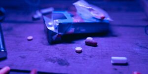 Pills scattered on dark wooden surface under purple lighting, with plastic bag visible in background.