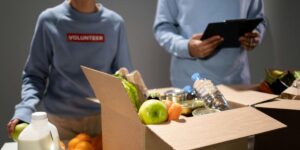 Volunteer in gray sweatshirt packing food boxes with fresh produce and groceries while colleague takes notes.