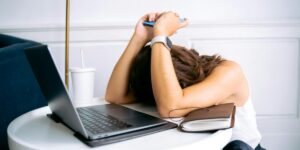 Woman lying face down on desk with laptop, appearing exhausted or frustrated. Smartwatch visible on wrist.