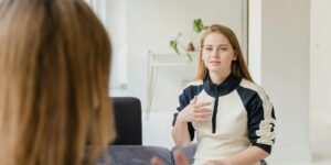 Young woman in black and white sweater gesturing while speaking, likely a sobriety coach in conversation with client in bright, minimalist room