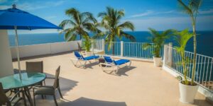 A view from a balcony overlooking a tropical beach. The balcony has blue lounge chairs and a blue umbrella. Palm trees and the bright blue ocean are visible in the background.