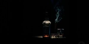 Bottle of alcohol and glass with smoking cigarette in a dimly lit room.