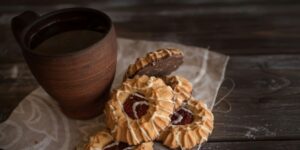 Traditional jam cookies and clay mug arranged on rustic wooden table with white cloth.