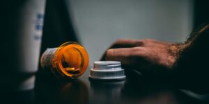 Orange prescription bottle and white cap in dim lighting with hand reaching toward medication