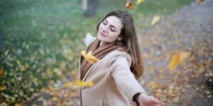 Woman in a beige coat enjoying the peaceful tranquility of a serene autumn forest, with her eyes closed and a serene expression on her face.