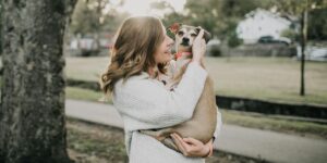 A smiling young woman warmly embracing her small dog in an outdoor park setting surrounded by trees and greenery.