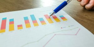 Blue pen pointing at colorful stacked bar chart and line graph on white paper