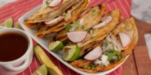 Crispy tacos garnished with radishes, limes, and cilantro served alongside consomé on red tablecloth