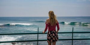 A woman standing on a balcony, looking out at the ocean. She is wearing a red top and a floral skirt.