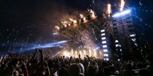 Massive concert crowd cheers as pyrotechnics and lights illuminate stage during energetic performance.