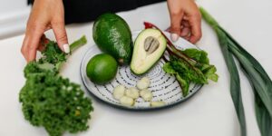 Hands preparing fresh ingredients including avocado, lime, kale, and garlic on patterned ceramic plate.