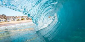 Massive turquoise wave curls over beach with oceanfront homes visible through crystal-clear water barrel.