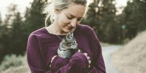 A young woman tenderly holds a small gray cat in her arms, surrounded by a serene outdoor forest environment