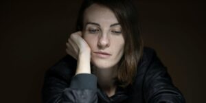 Young woman in dark jacket resting head on hand, looking downward in dim lighting