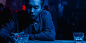 Person in blue lighting sitting at a bar with shot glasses, wearing a dark jacket