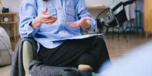 Person in light blue shirt gesturing while speaking, sitting in chair with coffee mugs on nearby table