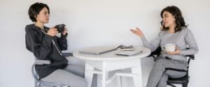 Two people having coffee and conversation at white table with laptop and notebooks