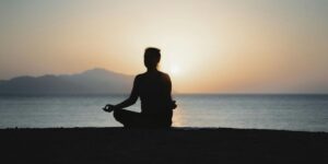 Silhouette of person meditating by ocean at sunset with mountains.